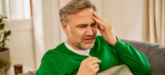 Mann in grünem Pullover sitzt auf einem Sofa und greift sich mit der linken Hand an die linke Stirn, da er aufgrund einer Erkältung Kopfschmerzen verspürt.
