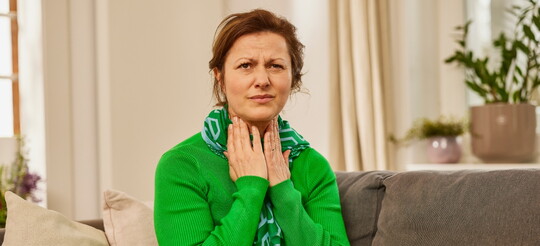 Frau auf der Couch mit Decke und Schal hält sich schmerzverzerrt die Hände an den Hals