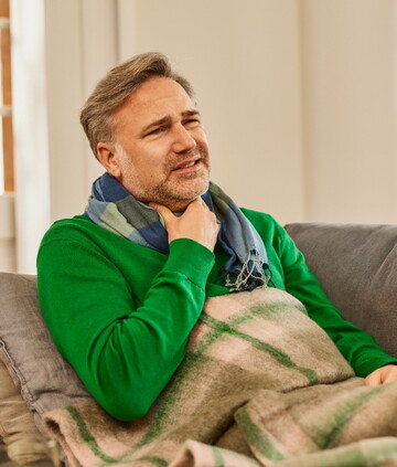 Mann mittleren Alters sitzt mit einer Decke auf der Couch und hält sich die Hand an den Hals, da er anscheinend Halsschmerzen hat
