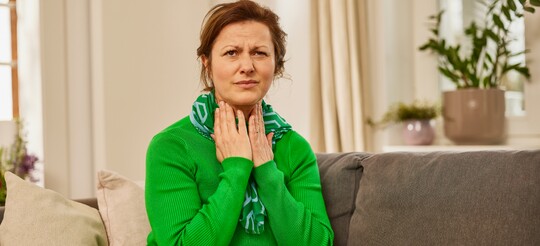 Frau mit Halsschmerzen sitzt eingekuschelt auf dem Sofa. Frau im grünen Pullover sitzt auf dem Sofa und hält sich mit den Händen den Hals, als ob sie Halsschmerzen verspürt. Sie trägt einen Schal und blickt mit einem Ausdruck des Unwohlseins. Die gemütliche Wohnzimmerumgebung mit Pflanzen und Decke vermittelt eine warme, ruhige Atmosphäre für Erholung und Selbstfürsorge.