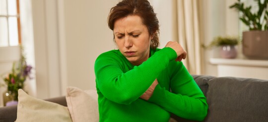 Frau hustet in die Armbeuge – mögliche Unterstützung bei Erkältung mit GeloProsed®. Frau in grünem Pullover sitzt auf einem Sofa und hustet in die Armbeuge, als Schutzmaßnahme gegen die Verbreitung von Keimen. Sie wirkt unwohl und möglicherweise erkältet. Die häusliche Umgebung mit Pflanzen im Hintergrund schafft eine beruhigende Atmosphäre, die zur Genesung beiträgt. GeloProsed® könnte bei der Linderung von Erkältungssymptomen hilfreich sein.