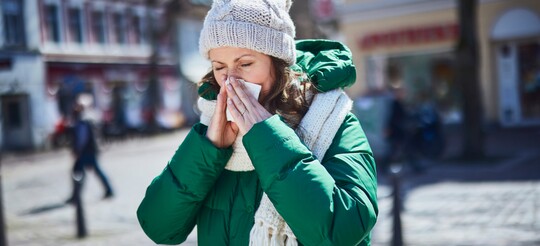 Frau in warmer Winterkleidung mit grüner Jacke, Mütze und Schal steht im Freien und putzt sich die Nase mit einem Taschentuch, während sie leicht fröstelt; Hintergrund zeigt unscharf eine städtische Szenerie.