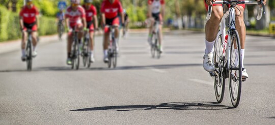 Radfahrergruppe bei sonnigem Training auf der Straße. Gruppe von Radfahrern in sportlicher Kleidung fährt auf einer breiten, sonnigen Straße. Die Radfahrer tragen rote Trikots und Helme und fahren eng beisammen, was auf ein gemeinsames Training oder ein Rennen hindeutet. Der Fokus liegt auf dem vorderen Radfahrer, während die anderen Teilnehmer leicht verschwommen im Hintergrund erscheinen. Die Umgebung ist grün und wirkt lebendig, passend zu einer aktiven, gesundheitsbewussten Lebensweise.