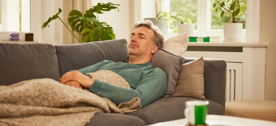 Ruhe und Entspannung im gemütlichen Wohnzimmer. Entspannter Mann, der auf einem Sofa liegt und ruht, zugedeckt mit einer Decke. Im Hintergrund sind grüne Pflanzen und ein Fenster, das Tageslicht hereinlässt, sichtbar. Auf dem Couchtisch stehen eine grüne Tasse und eine Schale.