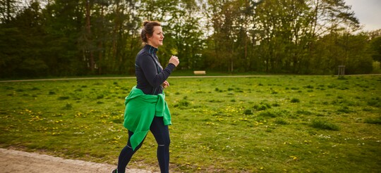 Frau beim Joggen im Park für Gesundheit und Wohlbefinden Frau joggt auf einem Weg in einem grünen Park, umgeben von Bäumen und Wiesen. Sie trägt sportliche Kleidung in dunklen Farben, mit einem grünen Pullover um die Taille gebunden, und bewegt sich dynamisch.