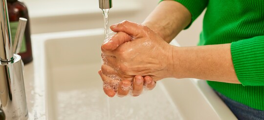 Hände waschen unter fließendem Wasser für gute Hygiene Nahaufnahme von Händen, die unter einem Wasserhahn mit fließendem Wasser gewaschen werden. Die Person trägt einen grünen Pullover und erzeugt mit Seife Schaum auf den Händen, während sie die Hygienepraktiken befolgt.