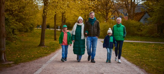 Familienausflug im herbstlichen Park Eine Familie spaziert an einem herbstlichen Tag auf einem Parkweg. Die Gruppe besteht aus zwei Erwachsenen, zwei Kindern und einer älteren Frau. Alle tragen warme Kleidung und Mützen, um sich vor der kühlen Jahreszeit zu schützen. Die gelb gefärbten Blätter der Bäume säumen den Weg und schaffen eine gemütliche Atmosphäre.