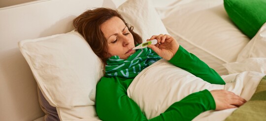 Frau liegt krank im Bett, eingehüllt in eine Decke und trägt einen Schal um den Hals. Sie hält ein Thermometer und überprüft ihre Temperatur, was auf Fieber oder Erkältungssymptome hindeutet. Die Szene im hellen Schlafzimmer mit weißen Kissen und grünen Akzenten vermittelt eine ruhige Atmosphäre für Erholung und Genesung.