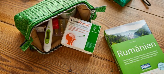 Offene grüne Reisetasche mit GeloProsed®-Packung, Fieberthermometer und weiteren Medikamenten, platziert auf einem Holztisch. Daneben liegt ein grüner Reiseführer für Rumänien und eine grüne Kamera, was auf eine Reisevorbereitung hindeutet. Die Szene betont die praktische Mitnahme von GeloProsed® zur Linderung von Erkältungssymptomen unterwegs.