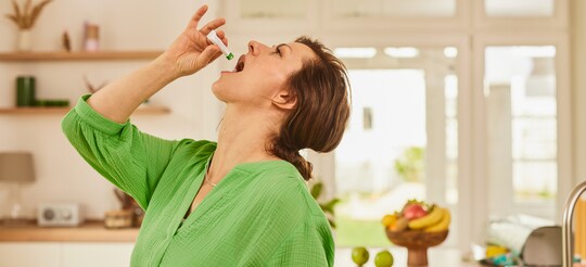 : Frau in grünem Oberteil nimmt in einem hellen, modernen Wohnzimmer GeloProsed® zur Linderung von Erkältungssymptomen ein. Sie hält eine kleine Flasche über ihren offenen Mund, um die Lösung einzunehmen. Im Hintergrund stehen frische Früchte und Dekorationsgegenstände, die eine warme und einladende Atmosphäre schaffen und die Bedeutung von Gesundheit und Wohlbefinden unterstreichen.