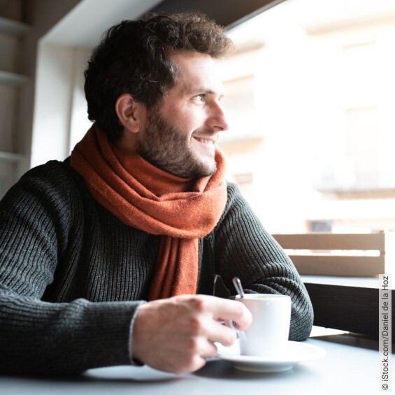 Glücklicher Mann mit Pullover und Schal, der eine Tasse heißen Getränk hält und aus dem Fenster schaut