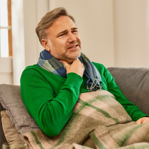 In der Anfangsphase einer Erkältung treten bei einem Mann Halsschmerzen auf. Er sitzt mit einer Decke auf der Couch und hält sich die Hand an den Hals