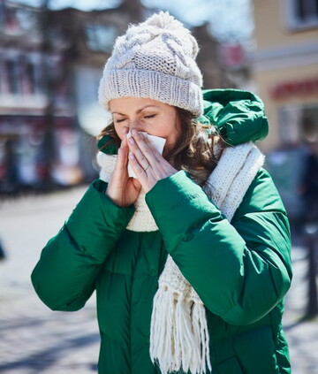 Frau mit grüner Winterjacke, Schal und Mütze steht draußen in der Stadt und schnäuzt sich die Nase. Sie ist sichtlich erkältet