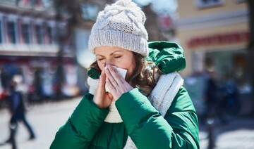Frau mit grüner Winterjacke, Schal und Mütze steht draußen in der Stadt und schnäuzt sich die Nase. Sie ist sichtlich erkältet