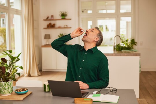 Mann nimmt GeloProsed® zur Linderung von Erkältungssymptomen ein. Mann mittleren Alters in grünem Hemd nimmt im Wohnzimmer GeloProsed® zur Linderung von Erkältungssymptomen ein. Er hält eine kleine Flasche über seinen offenen Mund, um die Lösung einzunehmen. Auf dem Tisch vor ihm stehen ein Laptop, ein Notizbuch und eine Brille, die eine entspannte häusliche Umgebung darstellen. Im Hintergrund sorgen Pflanzen und große Fenster für eine helle und freundliche Atmosphäre, passend zur gesundheitlichen Selbstfürsorge.