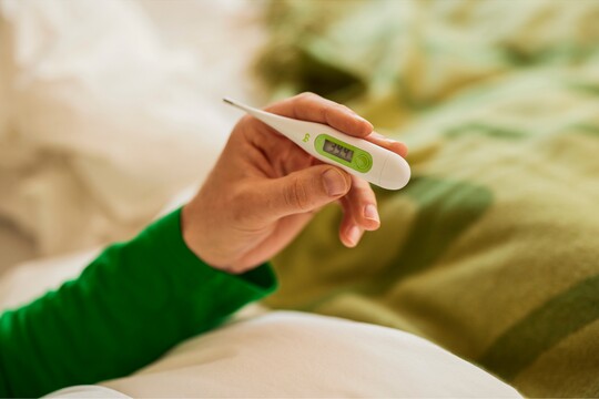 Fieberthermometer zeigt erhöhte Temperatur an. Nahaufnahme einer Hand, die ein digitales Fieberthermometer hält, das eine erhöhte Temperatur anzeigt. Im Hintergrund sind weiche, helle Bettdecken sichtbar, was auf eine gemütliche Umgebung zur Erholung bei Fieber hindeutet. Die Hand trägt einen grünen Ärmel, was zu einer warmen und fürsorglichen Atmosphäre beiträgt.