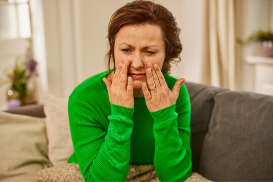Frau massiert Nebenhöhlen bei Erkältungsbeschwerden. Frau im grünen Pullover sitzt auf dem Sofa und massiert ihre Wangen unter den Augen, als ob sie Druck oder Schmerzen in den Nebenhöhlen verspürt. Ihr Gesichtsausdruck zeigt Unwohlsein, möglicherweise aufgrund von Erkältung oder Nebenhöhlenschmerzen. Die gemütliche Umgebung im Wohnzimmer vermittelt eine ruhige Atmosphäre für Erholung und Selbstfürsorge.