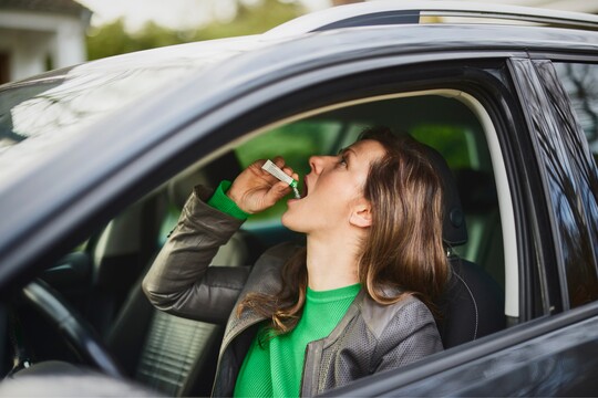 Eine Frau sitzt auf dem Fahrersitz eines Autos und nimmt GeloProsed® ein. Sie hält das Produkt in der Hand und hat den Kopf leicht nach hinten geneigt, um es einzunehmen. Sie trägt eine grüne Kleidung und eine Lederjacke, während sie sich im Auto befindet.