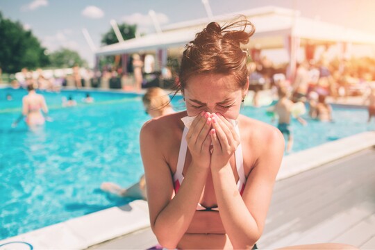 Junge Frau in Bikini sitzt am Beckenrand eines belebten Pools und schnäuzt sich die Nase, möglicherweise aufgrund einer Erkältung.