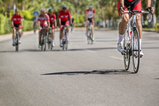 Radfahrergruppe bei sonnigem Training auf der Straße. Gruppe von Radfahrern in sportlicher Kleidung fährt auf einer breiten, sonnigen Straße. Die Radfahrer tragen rote Trikots und Helme und fahren eng beisammen, was auf ein gemeinsames Training oder ein Rennen hindeutet. Der Fokus liegt auf dem vorderen Radfahrer, während die anderen Teilnehmer leicht verschwommen im Hintergrund erscheinen. Die Umgebung ist grün und wirkt lebendig, passend zu einer aktiven, gesundheitsbewussten Lebensweise.