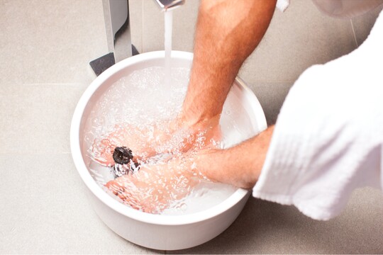 Fußbad zur Entspannung und Pflege. Füße einer Person in einem weißen Fußbad unter fließendem, warmem Wasser, umgeben von einer entspannten Atmosphäre und einem Bademantel. Die Szene betont die wohltuende Wirkung eines Fußbads zur Entspannung.