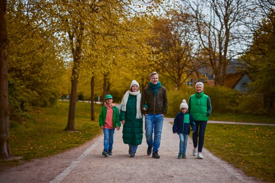 Eine Familie spaziert an einem herbstlichen Tag auf einem Parkweg. Die Gruppe besteht aus zwei Erwachsenen, zwei Kindern und einer älteren Frau. Alle tragen warme Kleidung und Mützen, um sich vor der kühlen Jahreszeit zu schützen. Die gelb gefärbten Blätter der Bäume säumen den Weg und schaffen eine gemütliche Atmosphäre.