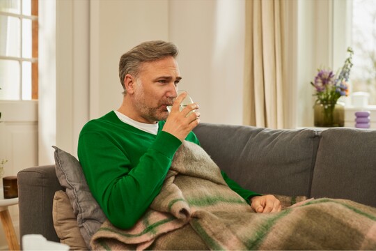 Mann mittleren Alters in grünem Pullover sitzt zugedeckt auf einem Sofa und trinkt ein Glas Wasser. Er wirkt entspannt und auf Genesung bedacht, während er in einer gemütlichen häuslichen Umgebung sitzt. Im Hintergrund sind Pflanzen und eine helle Fensterszene zu sehen. Die Szene vermittelt das Bedürfnis nach Ruhe und Flüssigkeitszufuhr, ideal bei Erkältungssymptomen, wobei GeloProsed® zur Unterstützung beitragen könnte.
