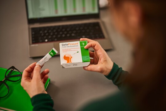 Nahaufnahme von zwei Händen, die eine Packung GeloProsed® und ein Sachet mit Pulver zum Einnehmen halten. Im Hintergrund steht ein geöffneter Laptop, daneben liegen eine grüne Mappe und eine schwarze Brille, was auf eine Arbeitsumgebung hindeutet. Die Szene vermittelt das praktische Einnehmen von GeloProsed® zur Linderung von Erkältungssymptomen, passend für den Alltag.