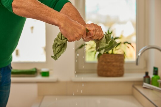 Nahaufnahme von Händen, die ein nasses grünes Tuch über einem Waschbecken auswringen, während Wasser heraustropft. Die Szene vermittelt den Eindruck einer vorbereitenden Maßnahme, möglicherweise für eine warme Kompresse zur Linderung von Beschwerden. Im Hintergrund sind Pflanzen und Badezimmerutensilien zu sehen, was eine ruhige, häusliche Atmosphäre betont, passend für Selbstpflege und Wohlbefinden.