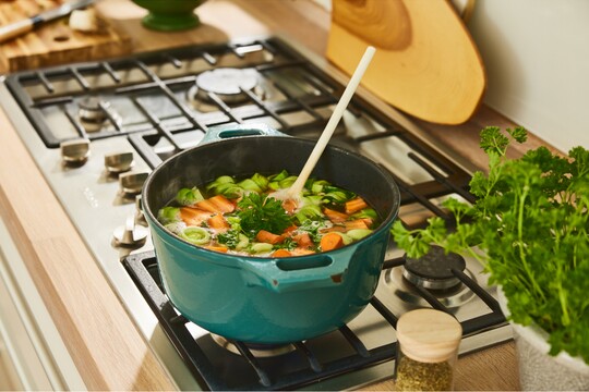 Ein blauer Topf mit frischer Gemüsesuppe, die auf einem Herd köchelt. Im Topf sind geschnittene Karotten, Lauch und Petersilie zu sehen, die zusammen eine gesunde, hausgemachte Suppe bilden. Ein hölzerner Kochlöffel ragt aus der Suppe. Daneben stehen frische Kräuter und Gewürze, was auf eine gesundheitsbewusste Zubereitung hinweist. Die Szene vermittelt Wärme und Wohlbefinden, passend als unterstützende Maßnahme bei Erkältungssymptomen.