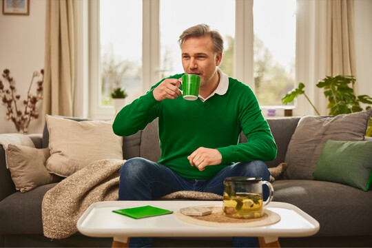 Mann in grünem Pullover sitzt entspannt auf einem Sofa und trinkt aus einer grünen Tasse. Vor ihm steht ein Tisch mit einer Teekanne voller Kräutertee und einem grünen Notizbuch. Die helle, gemütliche Wohnzimmerszene mit Pflanzen und Kissen im Hintergrund vermittelt Wärme und Entspannung, ideal für eine Erholungszeit, möglicherweise zur Unterstützung bei Erkältungssymptomen.