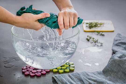 Hände wringen ein grünes Tuch über einer großen Glasschüssel mit Wasser aus, während Wasser heraustropft. Auf dem grauen Tisch sind frische Kräuter und ein Geschirrtuch zu sehen, die eine natürliche und beruhigende Atmosphäre schaffen. Die Szene vermittelt die Vorbereitung einer warmen Kompresse oder eines Kräuterumschlags zur Selbstpflege und Entspannung.