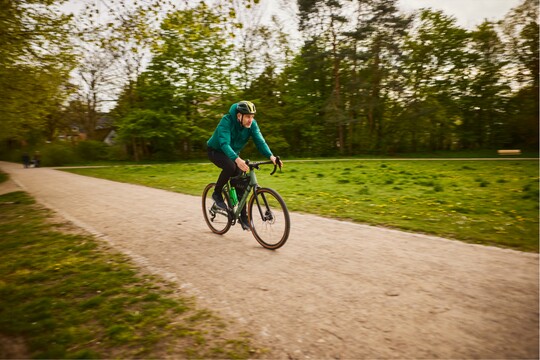 Mann beim Fahrradfahren im Grünen Mann in grüner Jacke und Helm fährt mit dem Fahrrad auf einem Waldweg entlang einer Wiese; sportliche Aktivität in der Natur.