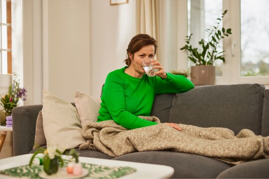 Frau in grünem Pullover, eingekuschelt in eine Decke auf einem grauen Sofa, trinkt ein Glas Wasser in einem hellen Wohnzimmer mit Pflanzen und gemütlicher Einrichtung, dargestellt als Teil der Erkältungserholung durch Ruhe und ausreichendes Trinken.