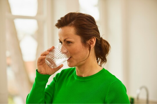 Frau in grünem Pullover trinkt ein Glas Wasser in einem hellen Raum mit Fenstern im Hintergrund.