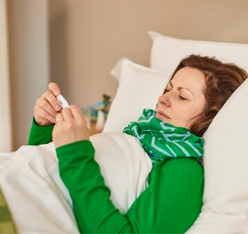 Frau liegt krank im Bett, eingehüllt in eine Decke und trägt einen Schal um den Hals. Sie hält ein Thermometer in der Hand und überprüft ihre Temperatur, was auf Fieber oder Erkältungssymptome hinweist. Die gemütliche Umgebung mit weichen Kissen und einem warmen Farbschema vermittelt Ruhe und Erholung.