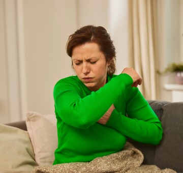 Frau in grünem Pullover sitzt auf einem Sofa und hustet in die Armbeuge, als Schutzmaßnahme gegen die Verbreitung von Keimen. Sie wirkt unwohl und möglicherweise erkältet. Die häusliche Umgebung mit Pflanzen im Hintergrund schafft eine beruhigende Atmosphäre, die zur Genesung beiträgt. GeloProsed® könnte bei der Linderung von Erkältungssymptomen hilfreich sein.