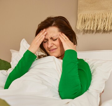 Frau liegt im Bett und hält sich mit schmerzverzerrtem Gesicht beide Hände an die Schläfen, möglicherweise wegen Kopfschmerzen oder Migräne. Sie trägt ein grünes Oberteil und ist in eine weiße Bettdecke gehüllt.