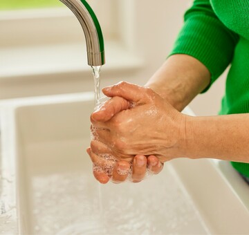 Nahaufnahme von Händen, die unter einem Wasserhahn mit fließendem Wasser gewaschen werden. Die Person trägt einen grünen Pullover und erzeugt mit Seife Schaum auf den Händen, während sie die Hygienepraktiken befolgt.