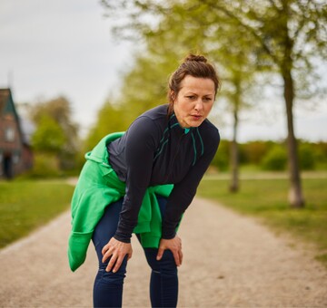 Frau in Sportkleidung steht auf einem Kiesweg in einer grünen Parklandschaft und beugt sich leicht nach vorne, als ob sie nach dem Joggen oder einer intensiven Trainingseinheit verschnauft. Sie trägt eine dunkelblaue Jacke und hat einen grünen Pullover um die Taille gebunden. Die Szene betont körperliche Aktivität und Erholung an der frischen Luft, passend für einen gesunden Lebensstil.