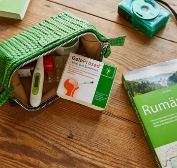 Offene grüne Reisetasche mit GeloProsed®-Packung, Fieberthermometer und weiteren Medikamenten, platziert auf einem Holztisch. Daneben liegt ein grüner Reiseführer für Rumänien und eine grüne Kamera, was auf eine Reisevorbereitung hindeutet. Die Szene betont die praktische Mitnahme von GeloProsed® zur Linderung von Erkältungssymptomen unterwegs.