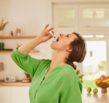 : Frau in grünem Oberteil nimmt in einem hellen, modernen Wohnzimmer GeloProsed® zur Linderung von Erkältungssymptomen ein. Sie hält eine kleine Flasche über ihren offenen Mund, um die Lösung einzunehmen. Im Hintergrund stehen frische Früchte und Dekorationsgegenstände, die eine warme und einladende Atmosphäre schaffen und die Bedeutung von Gesundheit und Wohlbefinden unterstreichen.