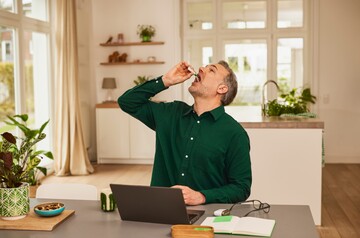 Mann mittleren Alters in grünem Hemd nimmt im Wohnzimmer GeloProsed® zur Linderung von Erkältungssymptomen ein. Er hält eine kleine Flasche über seinen offenen Mund, um die Lösung einzunehmen. Auf dem Tisch vor ihm stehen ein Laptop, ein Notizbuch und eine Brille, die eine entspannte häusliche Umgebung darstellen. Im Hintergrund sorgen Pflanzen und große Fenster für eine helle und freundliche Atmosphäre, passend zur gesundheitlichen Selbstfürsorge.