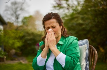 Frau im grünen Hemd sitzt draußen im Garten und hält sich ein Taschentuch vor die Nase, als ob sie niest oder Schnupfen hat. Ihr Gesichtsausdruck zeigt Unwohlsein, was auf Erkältungssymptome oder Allergien hinweist. Die grüne, naturreiche Umgebung betont eine natürliche Umgebung, in der sie sich möglicherweise ausruht und Genesung sucht. GeloProsed® könnte zur Linderung ihrer Symptome beitragen.