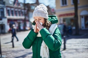 Frau in warmer Winterkleidung mit grüner Jacke, Mütze und Schal steht im Freien und putzt sich die Nase mit einem Taschentuch, während sie leicht fröstelt; Hintergrund zeigt unscharf eine städtische Szenerie.