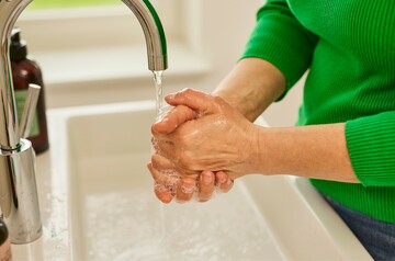 Nahaufnahme von Händen, die unter einem Wasserhahn mit fließendem Wasser gewaschen werden. Die Person trägt einen grünen Pullover und erzeugt mit Seife Schaum auf den Händen, während sie die Hygienepraktiken befolgt.