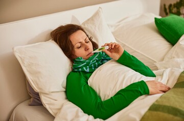Frau liegt krank im Bett, eingehüllt in eine Decke und trägt einen Schal um den Hals. Sie hält ein Thermometer und überprüft ihre Temperatur, was auf Fieber oder Erkältungssymptome hindeutet. Die Szene im hellen Schlafzimmer mit weißen Kissen und grünen Akzenten vermittelt eine ruhige Atmosphäre für Erholung und Genesung.