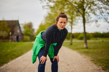Frau in Sportkleidung steht auf einem Kiesweg in einer grünen Parklandschaft und beugt sich leicht nach vorne, als ob sie nach dem Joggen oder einer intensiven Trainingseinheit verschnauft. Sie trägt eine dunkelblaue Jacke und hat einen grünen Pullover um die Taille gebunden. Die Szene betont körperliche Aktivität und Erholung an der frischen Luft, passend für einen gesunden Lebensstil.